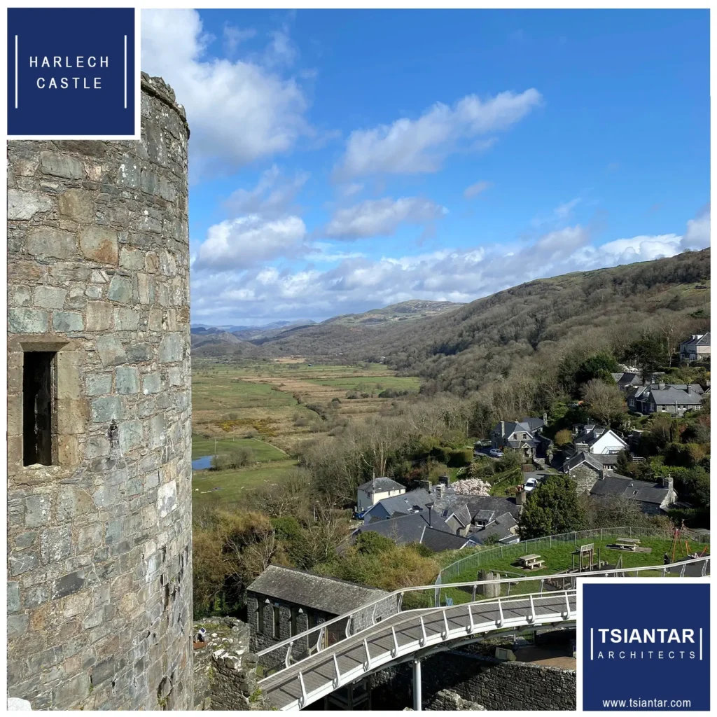 View from Harlech Castle overlooking a small village and verdant landscapes under a cloudy sky in North Wales, with the castle's stone wall and a logo of Tsiantar Architects in the corner.