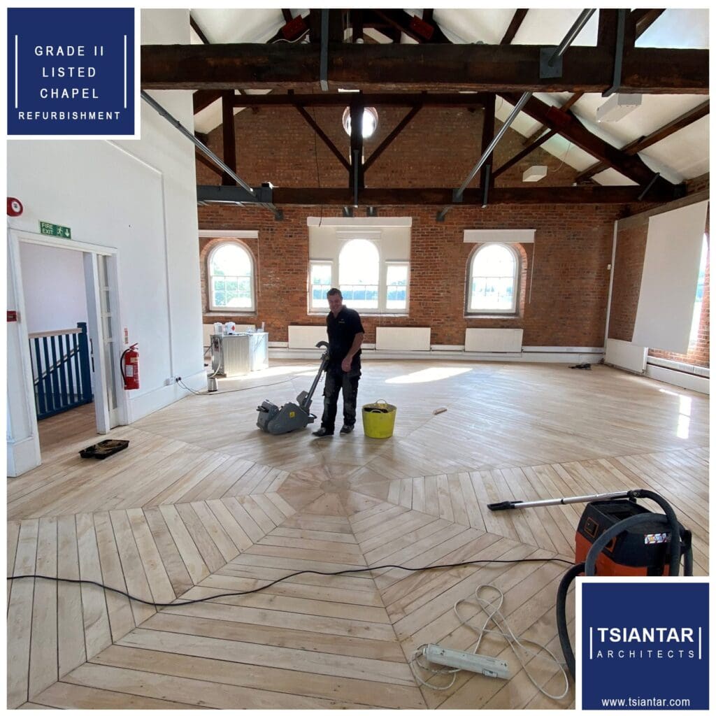 A worker skillfully sands the wooden floor in a beautifully refurbished chapel, where exposed brick walls and arched windows narrate stories of its past. The careful chapel refurbishment preserves both history and elegance.